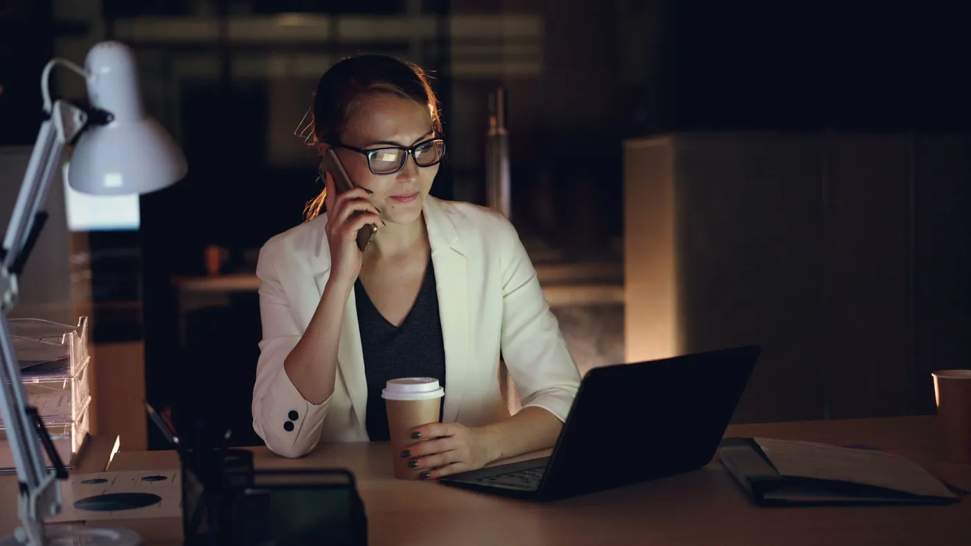 A woman in glasses on a phone call at her desk late at night, a laptop and coffee in front of her