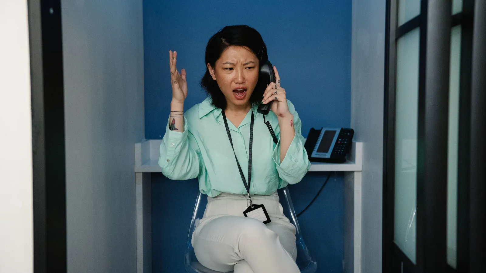 A woman in an office phone booth throwing up one hand in exasperation while talking on a desk phone